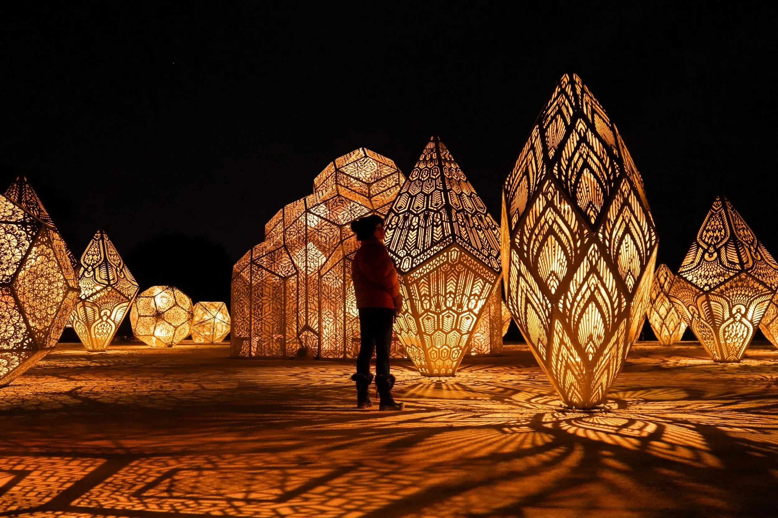 Illuminated faceted sculptures standing outdoors at night, their laser-cut patterns throwing intricate shadows across the ground as a visitor looks up at one