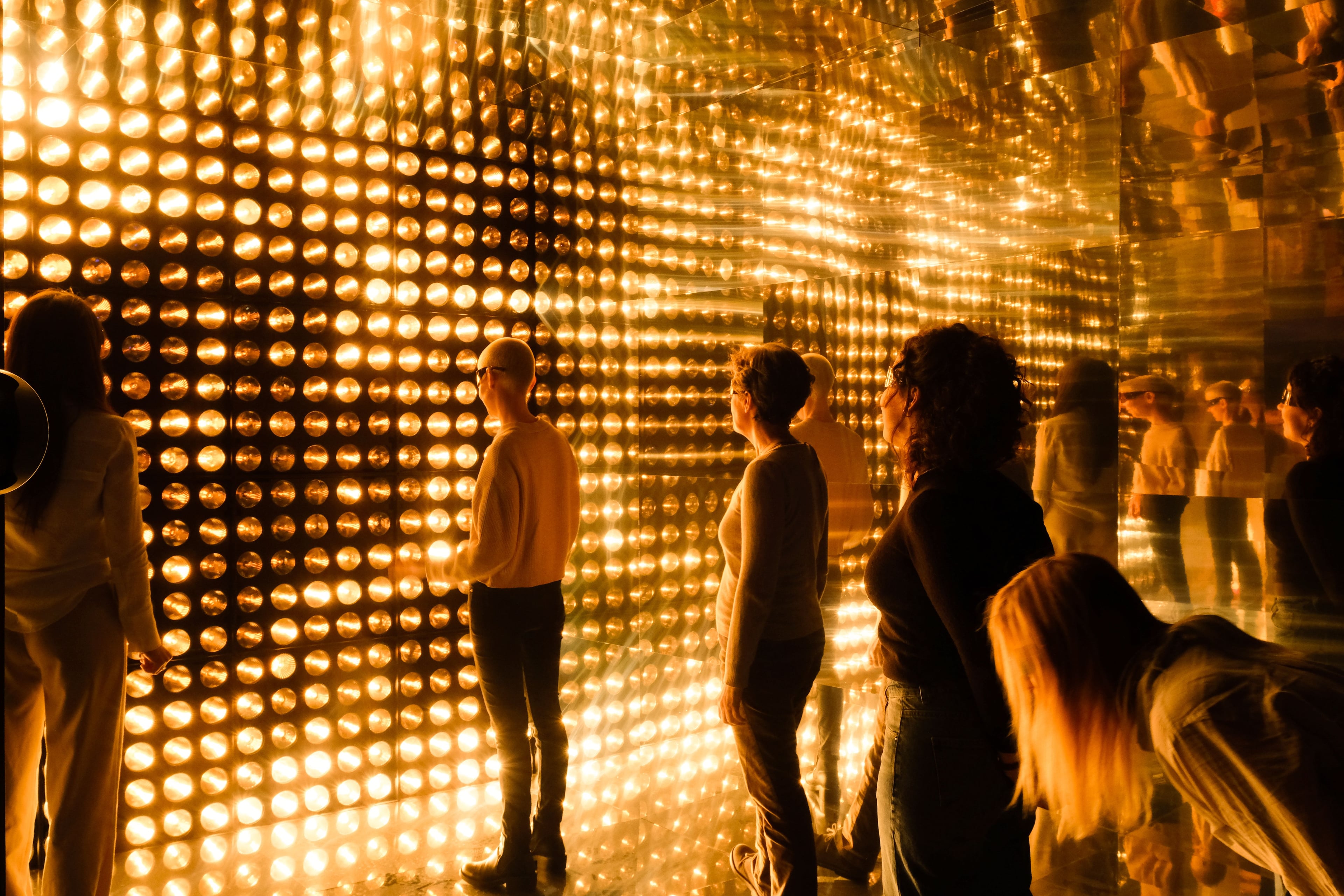 Visitors standing inside a mirrored room lined with dense grids of warm golden lights, their reflections repeating into the distance