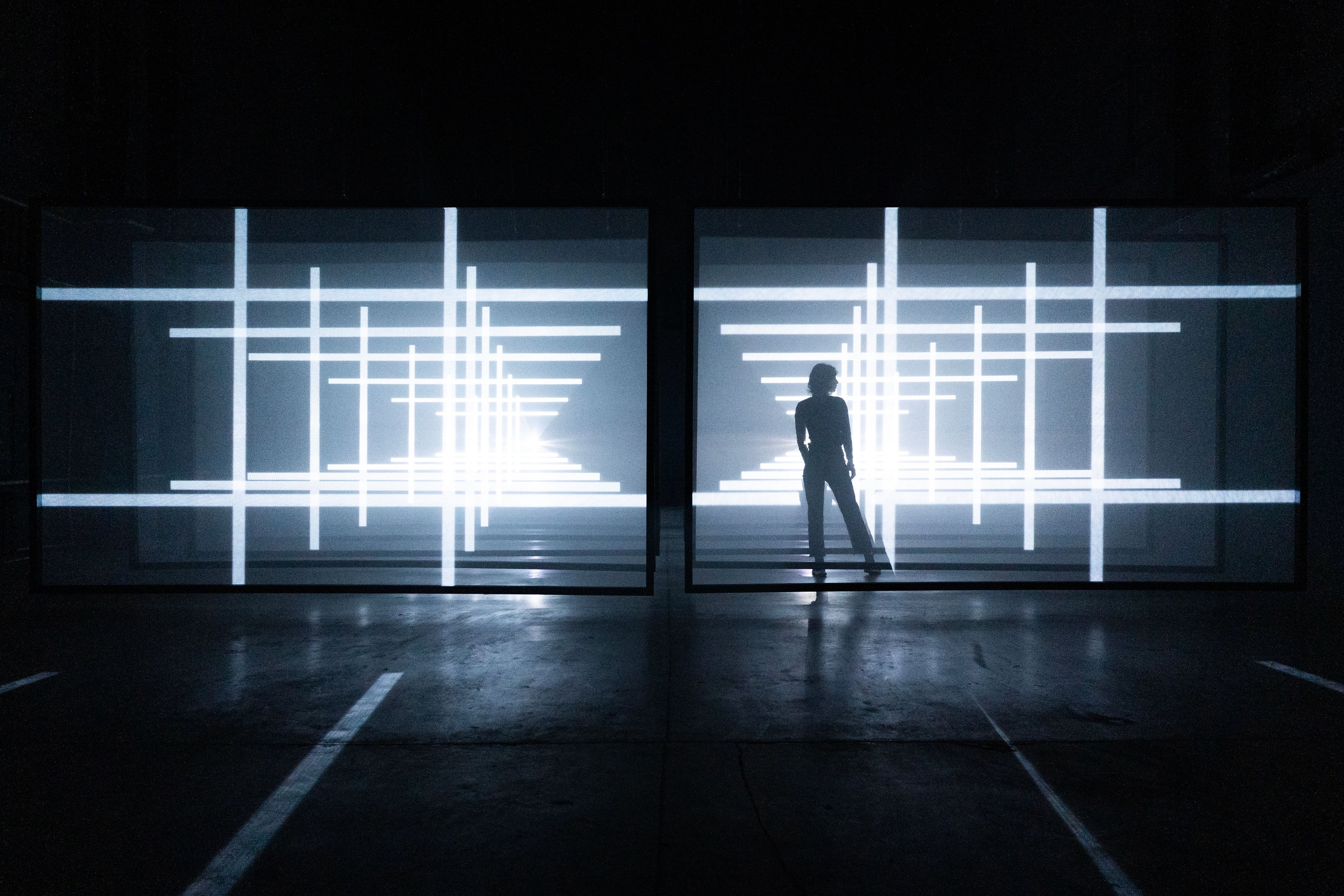 Two large screens of crossing white light bars facing each other across a dark concrete hall, with a figure standing silhouetted between them