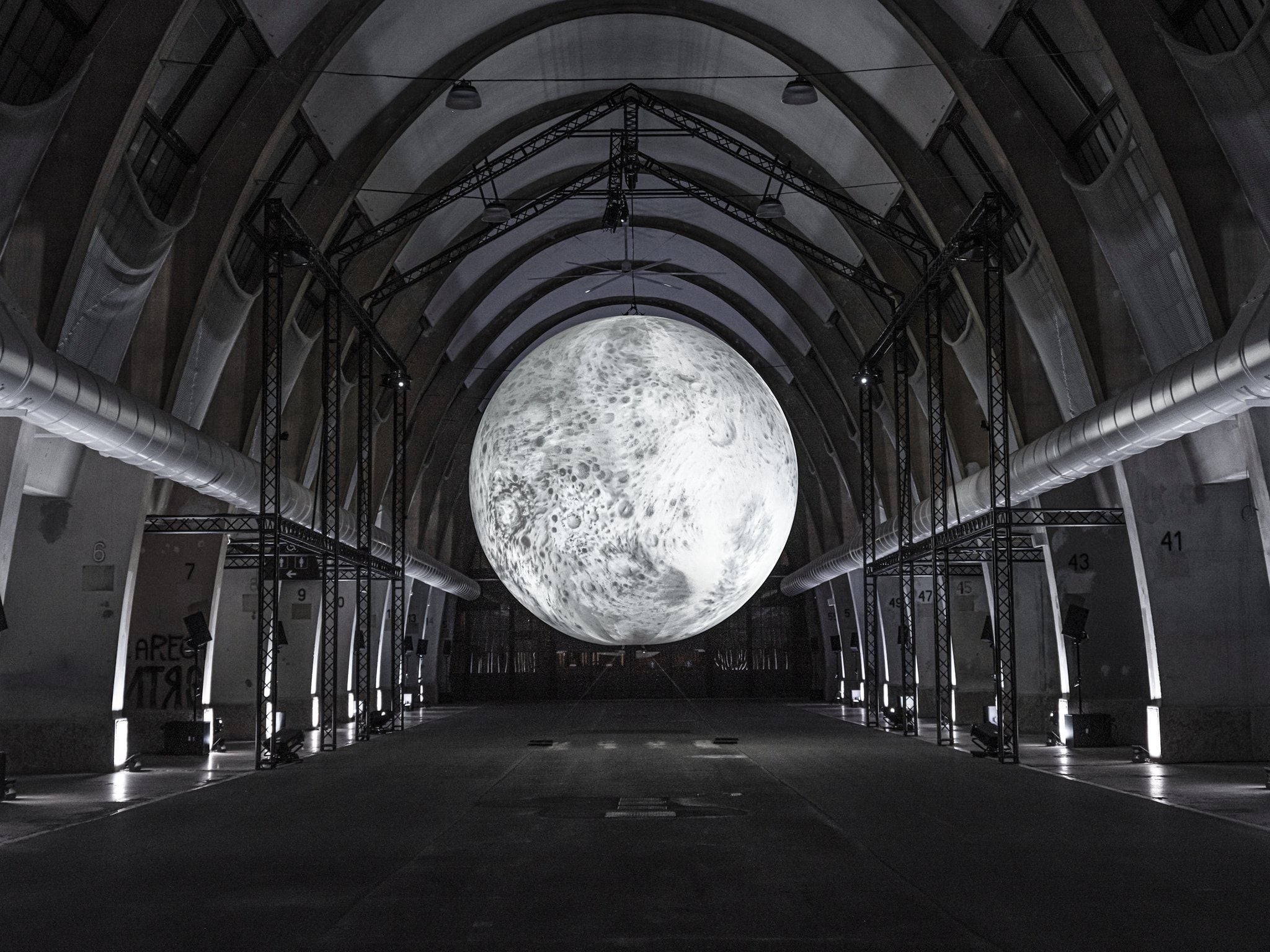 A vast illuminated sphere resembling the moon, suspended above the floor of a long vaulted industrial hall lined with lighting trusses